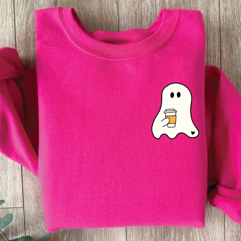 May include: A pink sweatshirt with a white ghost holding a cup of coffee. The ghost has two black eyes and a small heart on its side.