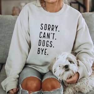 Może przedstawiać: Osoba w jasnoszarym swetrze z napisem "SORRY. CAN'T. DOGS. BYE." w kolorze czarnym. Biały pies siedzi na jej kolanach.