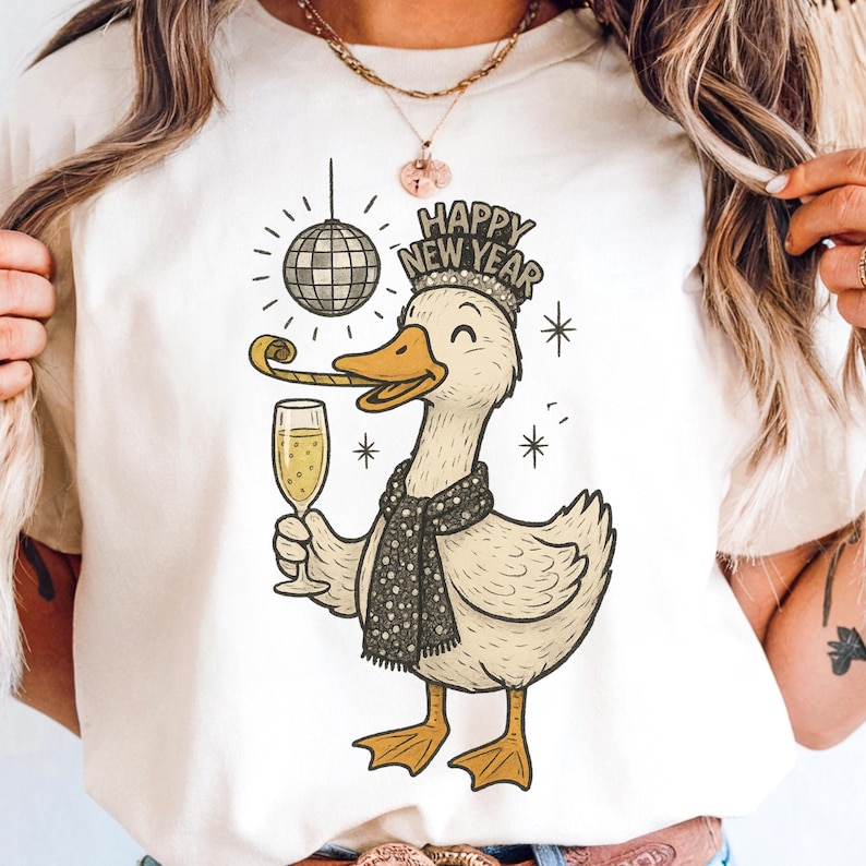 May include: Cream-colored t-shirt featuring a cartoon duck celebrating the New Year. The duck wears a crown and scarf, holding a champagne glass and party horn. The words "HAPPY NEW YEAR" are above the duck, with a disco ball and stars.