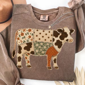 May include: A brown crewneck sweatshirt featuring a large patchwork cow graphic on the front. The cow design is composed of various fabric patterns including floral prints, polka dots, and brown cow spots. The inner neck label reads Comfort Colors.