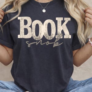 May include: Dark navy blue t-shirt with the text "BOOK snob" in a distressed, textured font. The word "BOOK" is in large capital letters, and "snob" is in a cursive font. The shirt is a casual style.
