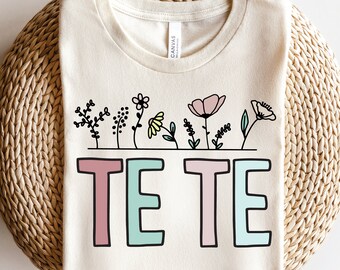 May include: A beige t-shirt featuring a floral design and the text "TE TE" in pink, green and blue.