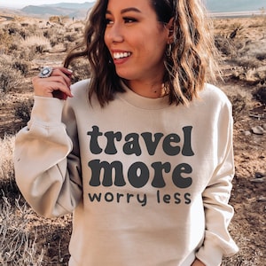 May include: A beige sweatshirt with the text "travel more worry less" in black retro-style lettering.