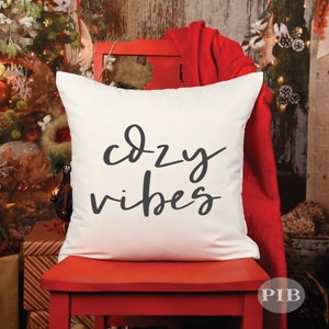 May include: White throw pillow with the text "cozy vibes" in black script font. The pillow is sitting on a red chair in front of a Christmas tree.