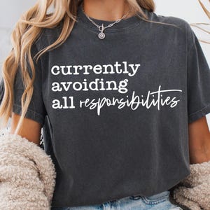May include: Dark gray t-shirt with the white text "currently avoiding all responsibilities." The shirt has a relaxed fit and short sleeves. The model is wearing a silver necklace and a cream-colored cardigan.