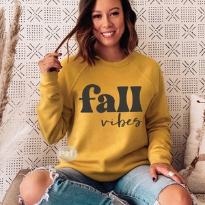 Może przedstawiać: Ż&oacute;łta bluza w kolorze musztardowym z napisem "fall vibes" czarnymi literami w blokach. Bluzę nosi osoba o brązowych włosach.