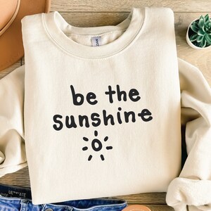 Op de afbeelding: Een crèmekleurig sweatshirt met de tekst "be the sunshine" in zwarte letters en een eenvoudige zonafbeelding.
