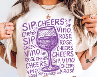 Op de afbeelding: Crèmekleurig T-shirt met een paarse wijnglasafbeelding en de woorden "Sip, Cheers, Vino, Rosé" in een herhalend patroon. Het ontwerp staat in het midden van de voorkant.