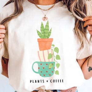 May include: Cream-colored t-shirt featuring a watercolor illustration of stacked plant pots and a mug. The top pot holds a green plant, the middle pot has pink stripes, and the bottom mug is teal with red hearts. The text "PLANTS + COFFEE" is below.