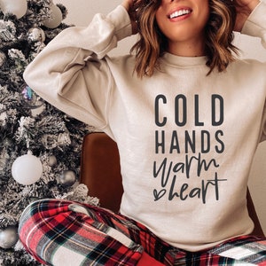 May include: A beige sweatshirt with the text "COLD HANDS warm heart" in black lettering. The sweatshirt is being worn by a person wearing red and black plaid pants.