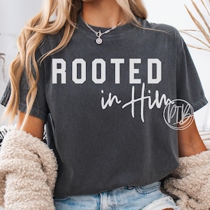 May include: Dark gray t-shirt with the words "ROOTED in Him" in white block and script lettering. The shirt is a casual style, with a relaxed fit. The text is centered on the front of the shirt.