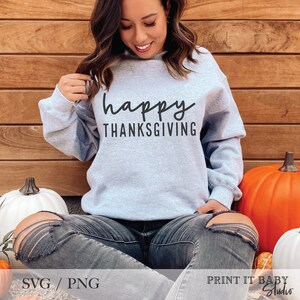 May include: A gray sweatshirt with the text "happy THANKSGIVING" in black lettering. The sweatshirt is being worn by a person sitting on a wooden surface with pumpkins.