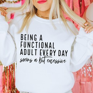 May include: White sweatshirt with the text "BEING A FUNCTIONAL ADULT EVERY DAY seems a bit excessive" in black. The sweatshirt is a crew neck style.