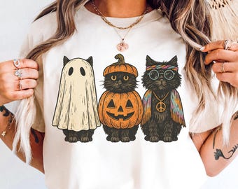 Könnte beinhalten: Beiges T-Shirt mit einem Halloween-Motiv von drei schwarzen Katzen. Eine ist als Geist verkleidet, eine andere als Kürbis und die dritte als Hippie mit einem Friedenssymbol.