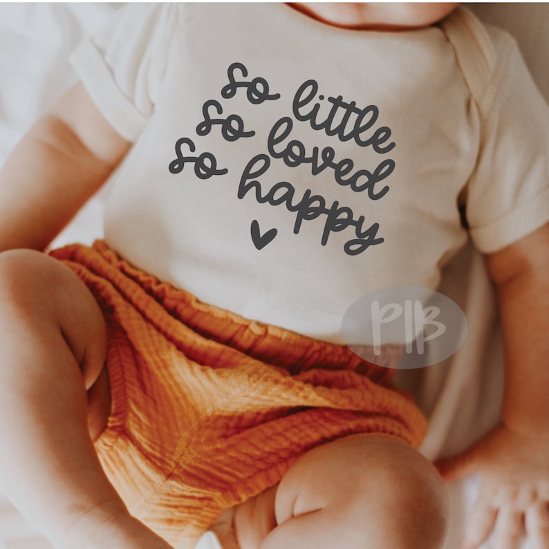 Puede incluir: Un body blanco para beb&eacute; con el texto "So little, so loved, so happy" en letra negra. Un peque&ntilde;o coraz&oacute;n est&aacute; debajo del texto. El body lo lleva un beb&eacute; con pantalones naranjas.
