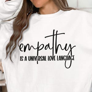Puede incluir: Sudadera blanca con texto negro que dice "la empatía es un lenguaje universal del amor".