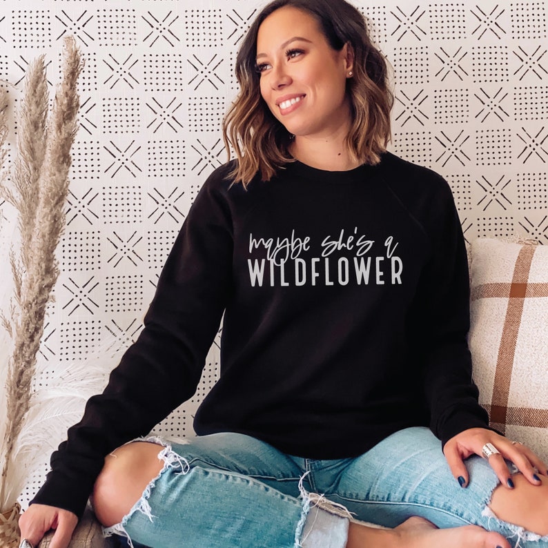 May include: A black sweatshirt with the text "maybe she's a wildflower" in white lettering.