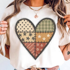 Op de afbeelding: Crèmekleurig T-shirt met een groot hartontwerp. Het hart is verdeeld in vier secties met verschillende patronen: stippen, ruitjes, bloemen en sterren. Het ontwerp heeft een gestikte omtrek.