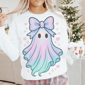 Puede incluir: Sudadera blanca con un dise&ntilde;o de fantasma pastel con lazo, corazones y estrellas. El fantasma tiene un degradado de rosa, azul y morado. El lazo es brillante. El dise&ntilde;o est&aacute; centrado en la parte delantera de la sudadera.