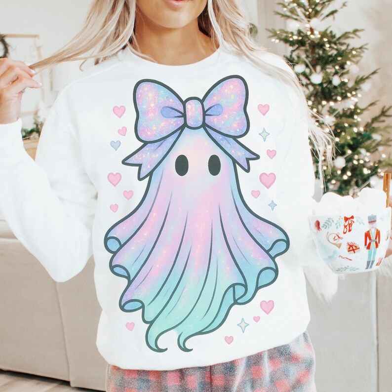 Puede incluir: Sudadera blanca con un dise&ntilde;o de fantasma pastel con lazo, corazones y estrellas. El fantasma tiene un degradado de rosa, azul y morado. El lazo es brillante. El dise&ntilde;o est&aacute; centrado en la parte delantera de la sudadera.