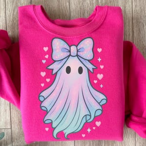 Puede incluir: Una sudadera rosa brillante con un dise&ntilde;o de fantasma de dibujos animados. El fantasma tiene un gran lazo azul claro brillante y un cuerpo azul claro y blanco. Peque&ntilde;os corazones y estrellas blancas rodean el fantasma.