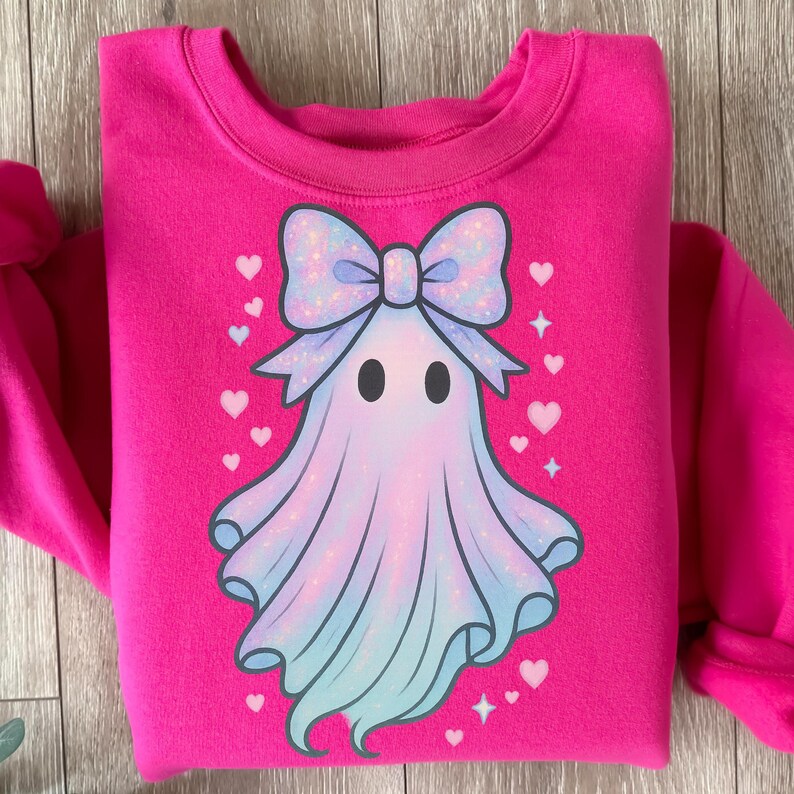 Puede incluir: Una sudadera rosa brillante con un dise&ntilde;o de fantasma de dibujos animados. El fantasma tiene un gran lazo azul claro brillante y un cuerpo azul claro y blanco. Peque&ntilde;os corazones y estrellas blancas rodean el fantasma.