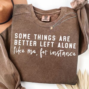 Puede incluir: Una sudadera marrón con el texto "SOME THINGS ARE BETTER LEFT ALONE like me, for instance" en blanco. Un collar plateado con un colgante está sobre la camisa. La camisa tiene una etiqueta Comfort Colors.