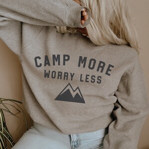 Peut inclure: Un sweat-shirt gris avec le texte "CAMP MORE WORRY LESS" et un graphique de montagne.