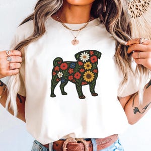 May include: Cream-colored t-shirt featuring a floral pug design. The pug silhouette is filled with red, white, yellow, and blue flowers. The shirt has a crew neck and short sleeves.