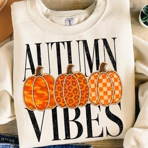 Op de afbeelding: Een crèmekleurige sweater met ronde hals, zwarte verweerde tekst "AUTUMN VIBES" en drie oranje pompoenen in het midden. De pompoenen hebben verschillende patronen: een camouflageprint, een luipaardprint en een ruitjespatroon.