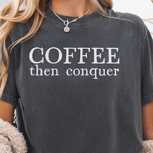 以下が含まれることがあります： 「COFFEE then conquer」の白い文字が入った濃いグレーのTシャツ。Tシャツは半袖でクルーネックです。ペンダント付きのシルバーのネックレスが見えます。
