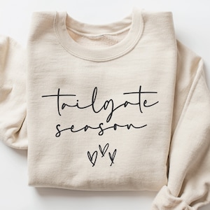 May include: A beige sweatshirt with the text "tailgate season" in black script and three black hearts below the text.