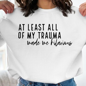 Peut inclure: Sweat-shirt blanc avec un texte noir qui dit "Au moins tout mon traumatisme m'a rendu hilarant".