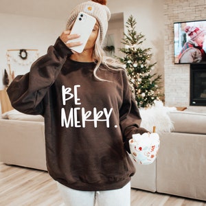 May include: A brown sweatshirt with the text "BE MERRY" in white lettering. The sweatshirt is worn by a person holding a mug of hot chocolate with marshmallows.