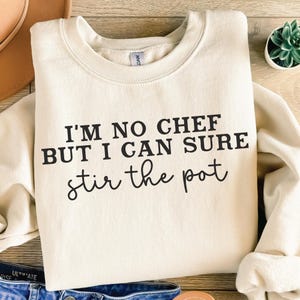 Op de afbeelding: Een crèmekleurig sweatshirt met de tekst "I'm no chef but I can sure stir the pot" in zwart gedrukt.