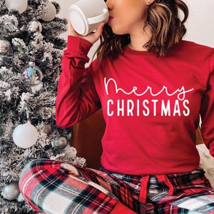 May include: A red long-sleeved shirt with the text "merry christmas" in white script. The person is wearing red and black plaid pajama pants.