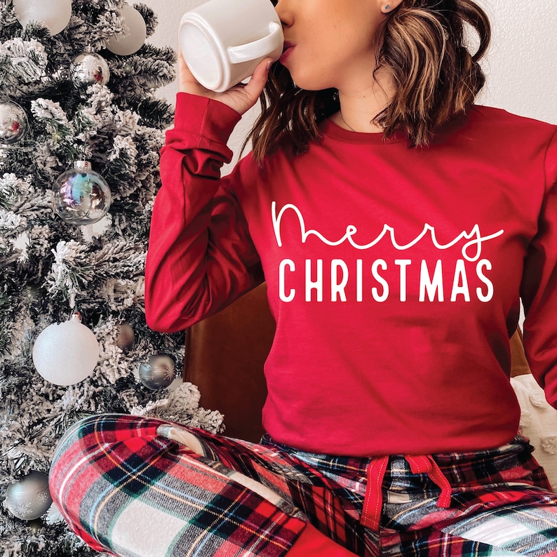 May include: A red long-sleeved shirt with the text "merry christmas" in white script. The person is wearing red and black plaid pajama pants.