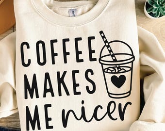 May include: Cream-colored sweatshirt with the text "COFFEE MAKES ME nicer" in black. A black and white graphic of a coffee drink with a heart is on the right side. The sweatshirt is folded on a wooden surface.