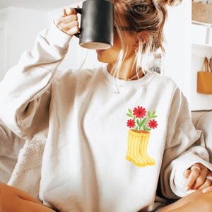 May include: Cream-colored sweatshirt featuring a graphic of red flowers and green leaves in a yellow rain boot. The sweatshirt has a crew neck and long sleeves. A person is holding a black mug.