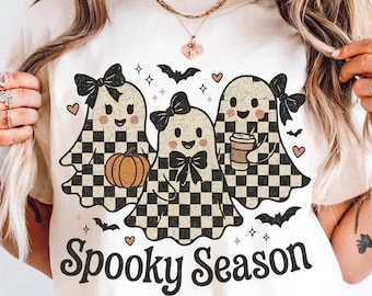 May include: Cream-colored t-shirt featuring three cartoon ghosts with checkered patterns, bows, and smiling faces. One ghost holds a pumpkin, another a coffee cup. The text "Spooky Season" is printed below the ghosts, with bats and hearts.