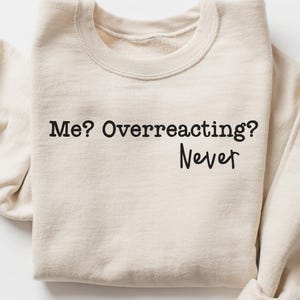 Peut inclure: Un sweat-shirt beige avec le texte "Me? Overreacting? Never" imprimé en noir. Le sweat-shirt à col rond est plié, mettant en valeur le motif sur le devant. Le haut décontracté est en matière douce.