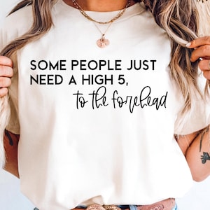 May include: Cream-colored t-shirt with the text "SOME PEOPLE JUST NEED A HIGH 5, to the forehead" in black. The shirt is a casual style, suitable for everyday wear.
