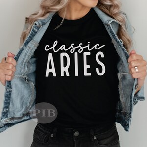 Puede incluir: Una camiseta negra con texto blanco que dice "classic ARIES". La camiseta la lleva puesta una persona con una chaqueta vaquera.