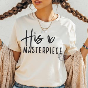 Puede incluir: Camiseta color crema con la frase "His Masterpiece" en escritura negra. El diseño incluye un símbolo de corazón. La camiseta tiene cuello redondo y mangas cortas, y está hecha de un material suave.