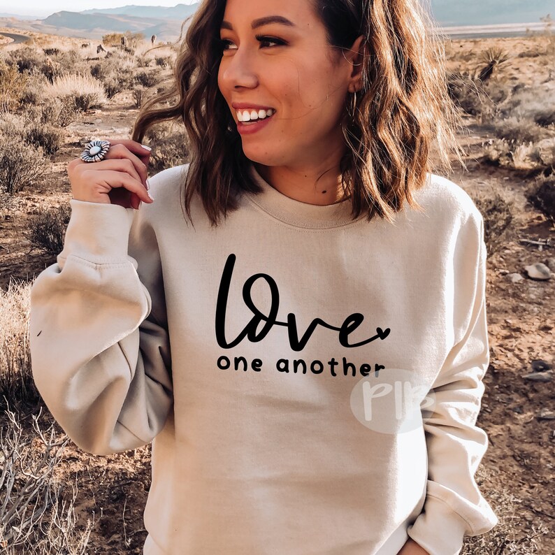 May include: Beige sweatshirt with the words "Love one another" in black script. The sweatshirt has long sleeves and a crew neck. The background shows a desert landscape with mountains and dry brush.