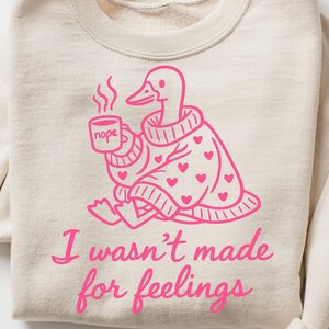 May include: Beige sweatshirt with a pink graphic of a duck wearing a sweater and holding a mug that says "nope." The duck is surrounded by pink hearts. The text "I wasn't made for feelings" is below the duck.