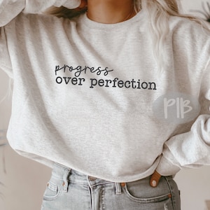 May include: A light gray sweatshirt with the words "progress over perfection" printed in black script. The sweatshirt is a crew neck style and appears to be made of a soft material. The model is wearing blue jeans.