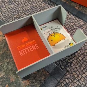 May include: A grey card game organiser with two compartments. One compartment holds a deck of Exploding Kittens cards, the other holds a single card with a taco cat illustration and the text "I am a palindrome!"