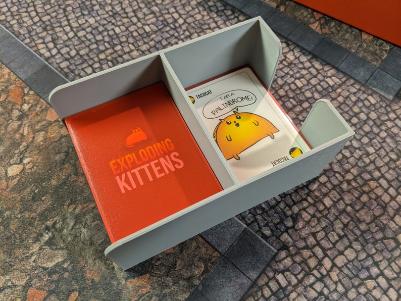 May include: A grey card game organiser with two compartments. One compartment holds a deck of Exploding Kittens cards, the other holds a single card with a taco cat illustration and the text "I am a palindrome!"