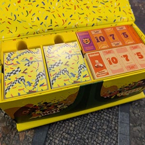 May include: A yellow box with a lid, filled with cards and game pieces. The box has a confetti design on the inside and outside. The cards have a checkered flag design and the game pieces have the numbers 10, 7, and 5 printed on them.